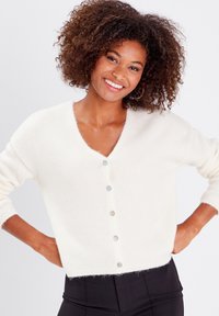 Cardigan blanc court avec un décolleté en V, des manches longues et une fermeture à cinq boutons ronds. Texture douce et pelucheuse avec une coupe ample et un ourlet côtelé.
