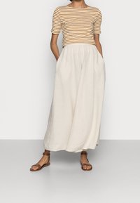 Gestreepte top met korte mouwen in bruin en crèmekleur, gecombineerd met een lange, vloeiende crèmekleurige rok. Bruine platte sandalen zorgen voor een contrasterend element.