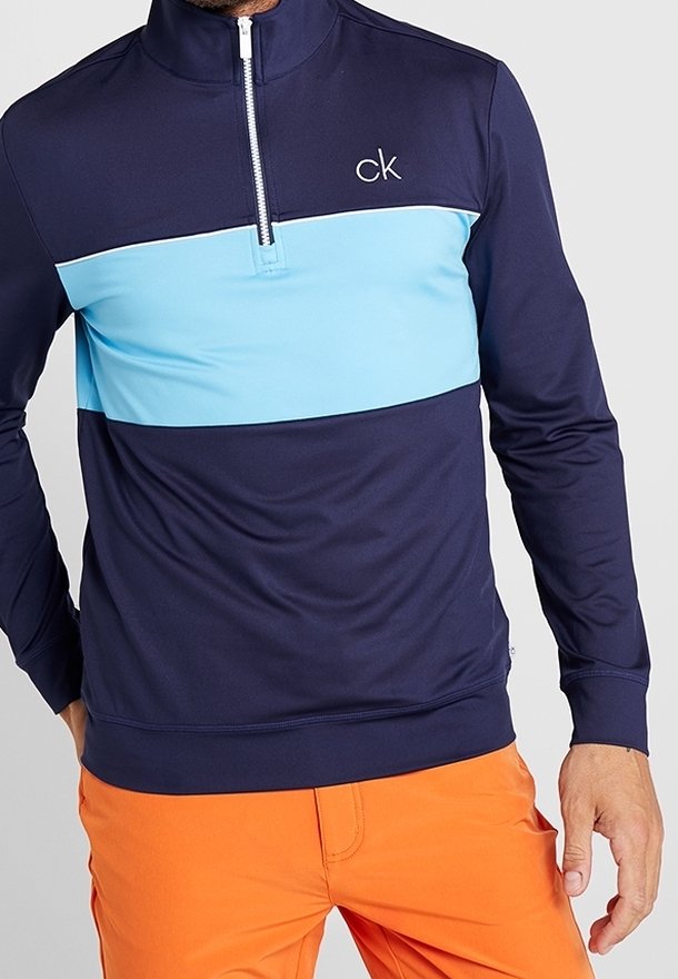 Námorně modrý svetr s dlouhým rukávem a polovičním zipem, světle modrý horizontální pruh, logo CK na hrudi, měkký a pružný materiál. Oranžové kalhoty.