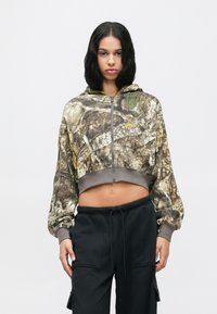 Ung kvinde står op, iført en cropped camouflage zip-up hoodie og sorte cargo bukser foran en ensfarvet hvid baggrund.