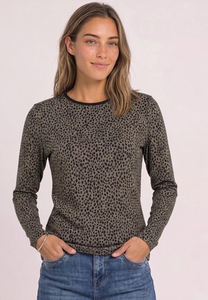 Femme aux cheveux châtain clair portant un haut vert olive à manches longues avec des taches noires et un jean bleu, debout les mains dans les poches.