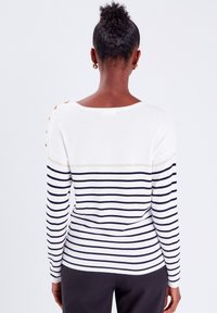 Pull rayé blanc et marine avec des manches longues, une coupe décontractée et des boutons aux épaules. Texture tricotée douce avec un large décolleté.