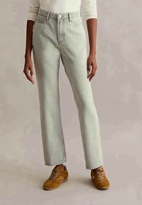 Osoba oblečená v svetlošedých vysokých džínsach, bielom tričku s dlhým rukávom a hnedých šnúrkových teniskách, stojí proti neutrálnej kulise.