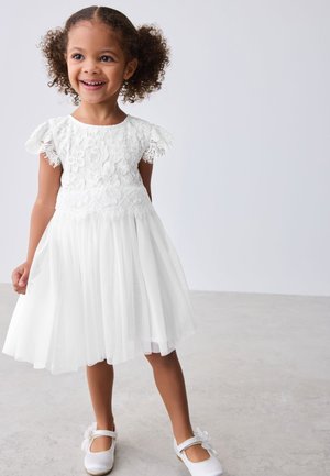 Lachend jong meisje met krullend haar, gekleed in een witte kanten jurk en witte schoenen, staand op een lichtgrijze vloer tegen een effen achtergrond.