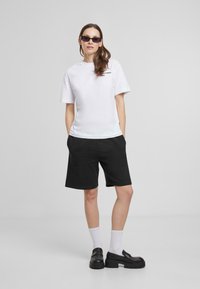 Hvit bomull T-skjorte med grafisk tekst, kombinert med svarte bomullsshorts. Modellen har på seg svarte platåsko og hvite ankelsokker.