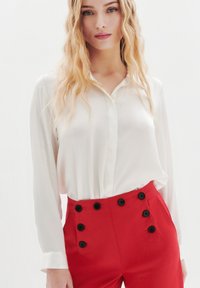 Vit blus med lång ärm och knappad krage, parat med högmidjade röda byxor med svarta dekorativa knappar på sidan.