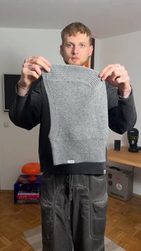 Indumento grigio lavorato a maglia con costine testurizzate, forma rettangolare ed elementi di design curvi. Presenta una piccola etichetta del marchio all'orlo.