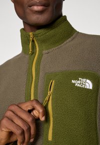 Giacca in fleece verde e marrone, con un colletto alto, zip gialla e una tasca sul petto con il logo The North Face in bianco.