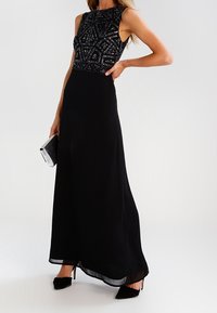 Kvinna håller en silverclutch och bär en ärmlös svart klänning med pärlbroderad bodice och lång flödande kjol, ihop med svarta klackar.