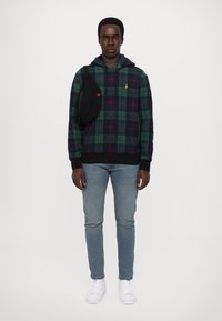 Felpa a quadri verdi in tessuto morbido, con pannello nero sui lati, abbinata a jeans azzurri chiari e sneakers bianche.