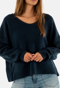 Maglione lavorato a maglia blu navy con scollatura a V, vestibilità oversize, orlo e polsini a coste. Il materiale sembra morbido e texturizzato, offrendo calore.