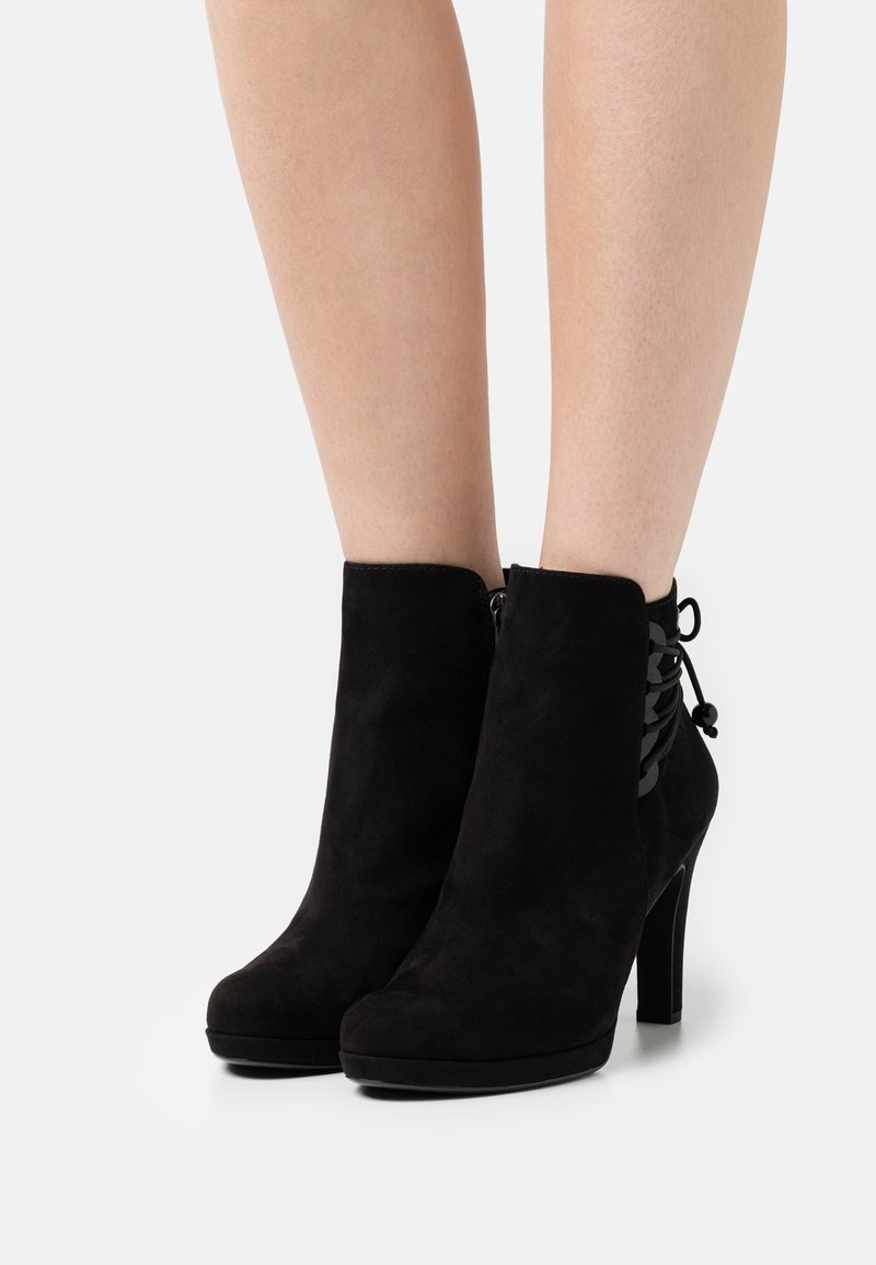 Bottes courtes en daim noir avec talons hauts et détails à lacets à l'arrière portées sur des jambes nues sur un fond uni.