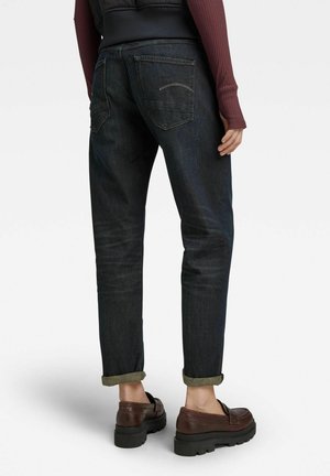 Person, die dunkle, hochgekrempelte Jeans und braune Leder-Loafers trägt, steht mit entspannten Händen an den Seiten vor weißem Hintergrund.