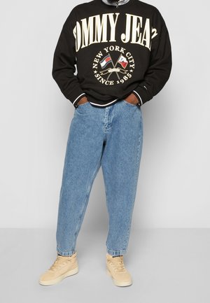 Personne portant un sweat-shirt noir Tommy Jeans avec une impression de New York City, un jean bleu clair et des baskets montantes beiges, les mains dans les poches.