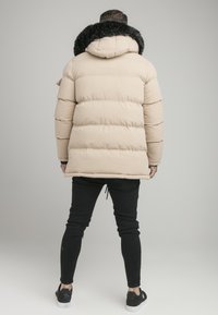 Helle beige Daunenjacke mit einer schwarzen Kunstfellkapuze, die horizontale Steppnähte aufweist. Kombiniert mit schwarzen Slim-Fit-Jeans und schwarzen Schuhen.