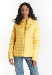 Gul puffjacka med horisontellt quiltat mönster, dragkedja framtill och hög krage. Kombinerad med blå denimbyxor.