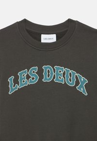 Σκούρο γκρι φούτερ με στρογγυλή λαιμόκοψη. Διαθέτει το κείμενο "LES DEUX" σε ανοιχτό μπλε χρώμα, σε στρωμένο σχέδιο, με απαλό, ελαφρώς υφασμένο ύφασμα.