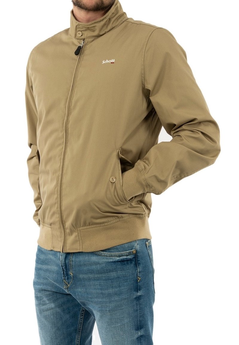 Veste bomber beige avec un col montant, fermeture éclair à l'avant et poignets élastiques. Présente des poches latérales et un logo "Schott" en rouge. Un jean en denim est porté en dessous.