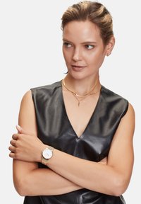 Montre avec bracelet en maille dorée et cadran blanc, portée sur un avant-bras nu. L'arrière-plan présente un haut en cuir noir lisse avec un décolleté en V profond.