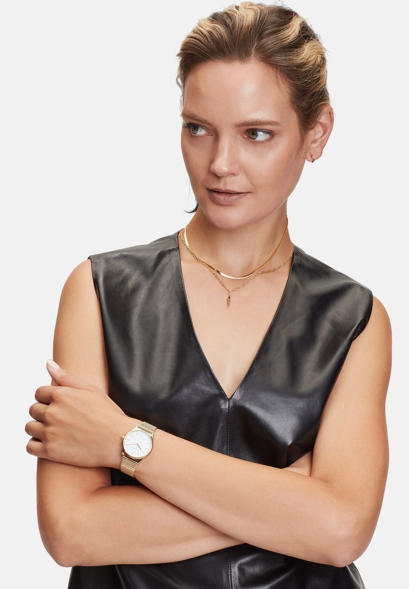 Montre avec bracelet en maille dorée et cadran blanc, portée sur un avant-bras nu. L'arrière-plan présente un haut en cuir noir lisse avec un décolleté en V profond.