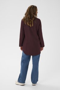 Donna con lunghi capelli mossi, veste una tunica a maniche lunghe color bordeaux scuro e jeans blu larghi, in piedi di spalle su uno sfondo bianco.