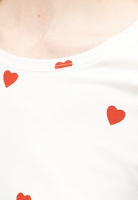 Tissu blanc parsemé de formes de cœurs rouges près d'un col, montrant une partie du haut de la poitrine et de la clavicule d'une personne.