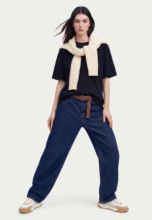 Donna in piedi con le gambe divaricate, indossa una t-shirt nera oversize, jeans blu scuro, un maglione beige di lana posato sulle spalle e scarpe da ginnastica bianche.