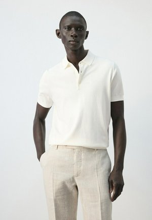 Hombre vistiendo una camisa tipo polo de manga corta blanca y pantalones de lino color beige claro sobre un fondo claro y liso.