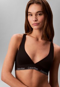 Czarny bezprzewodowy stanik o głębokim dekolcie w kształcie V, wykonany z gładkiego materiału. Posiada kontrastowe białe logo na pasku. Styl minimalistyczny.