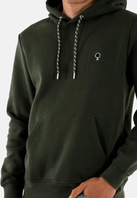Sudadera verde oscuro hecha de tela suave, con un bolsillo frontal, cordones ajustables con puntas metálicas y un pequeño logo de árbol en el pecho.