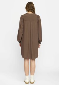 Rochie maro cu mâneci lungi, cu o croială lejeră, design pliat în spate și detalii texturate la umeri, asociată cu încălțăminte albă până la gleznă.