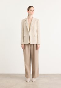 Beige rakenne bleiseri, jossa on lovettu kaulus, yhdistettynä löysiin beigeihin housuihin ja vaaleisiin korkokenkiin. Sileä, kudottu kangas; minimalistinen muotoilu.