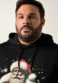 Schwarzer Kapuzenpullover mit einem grafischen Druck von Santa Claus, der einen rot-weißen Hut trägt, umgeben von Schneeflocken und mit einem strukturierten Bart.