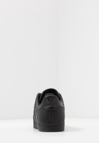 Musta Adidas-tossu, takanäkymä, jossa on tunnusomainen kolmilehtinen logo kantapäässä, asetettuna minimalistiselle valkoiselle taustalle.