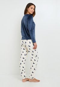 Top in velluto blu navy a maniche lunghe abbinato a pantaloni crema con silhouette di gatti blu scuro. Tessuto morbido, design aderente e stile casual.