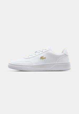Hvit lær sneakers med en glatt tekstur, flat gummisåle og en gulllogoaccent på siden. Har tradisjonell snøring.