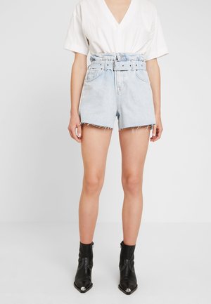 Femme portant un short en jean bleu clair taille haute avec une ceinture, un haut blanc à col en V et manches courtes, et des bottines noires avec embouts métalliques.