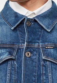 Närbild av en blå denimjacka med synliga knappar och bröstfickor, som bärs över en vit skjorta med krage.