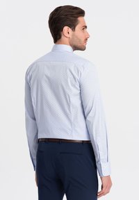 Camicia a maniche lunghe di un blu chiaro con piccole pois blu scuro, con colletto standard e polsini con bottone, indossata con pantaloni blu navy.