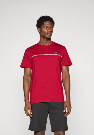 T-shirt en coton rouge avec un col rond, des manches courtes et une rayure blanche contrastante sur la poitrine. Logo Lacoste sur le côté gauche.