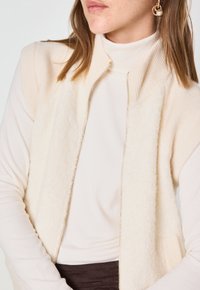 Gilet senza maniche color crema con una parte frontale morbida e texturizzata, bordi a coste e chiusura con zip. Indossato sopra un top a dolcevita leggero.