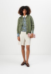 Donna che indossa un cardigan di maglia verde, una maglietta blu a fantasia, pantaloni corti beige, mocassini marroni e tiene una borsa a mano rosa su uno sfondo bianco.