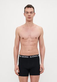 Boxers noirs à coupe ajustée, avec une ceinture élastique blanche à logo. Le modèle est torse nu, mettant en valeur un fond neutre.