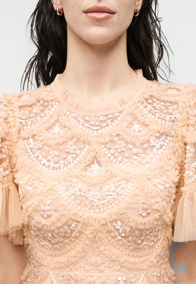 Γυναίκα με διαφανές ροδακινί δαντελένιο τοπ με φλοράλ και κυματιστά παγιετέ μοτίβα και κοντά βολάν μανίκια, απεικονιζόμενη από τους ώμους μέχρι τα χείλη.