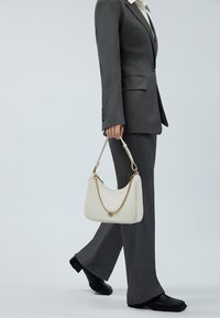 Vit läderhandväska med en böjd form, utrustad med en guldkedja och ett handtag ovanpå. Modellen bär en grå dräkt med utställda byxor.