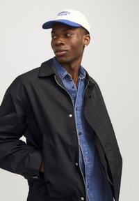 Giacca nera con zip frontale, abbinata a una camicia di denim blu. Cappellino bianco con visiera blu, con del testo. Il modello guarda di lato.