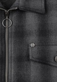 Veste en laine grise avec une finition texturée, fermeture éclair argentée et un accent en forme de rose sur la poche poitrine. Présente un design moderne et structuré.