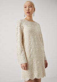 Robe en dentelle crème avec des motifs découpés circulaires, manches longues et ourlet festonné. Texture lisse, tissu léger.