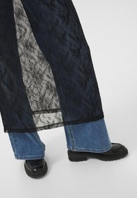 Long black lace overlay with a floral pattern, layered over blue wide-leg jeans and black platform shoes. Textured and sheer details visible.