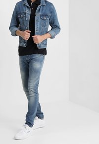 Džínová bunda se dvěma náprsními kapsami, černé polo tričko, světle modré slim-fit džíny a bílé tenisky. Casual outfit s vrstvenými texturami.